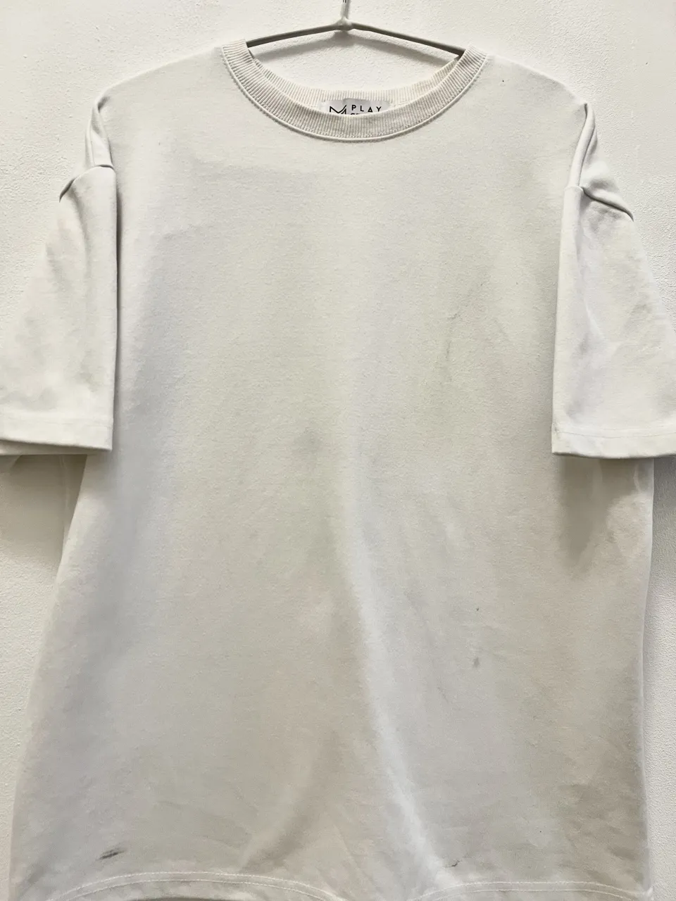 Remera de algodón color crudo, con cuello redondo acanalado y mangas cortas. Presenta algunas manchas pequeñas y detalles de uso en la parte inferior.