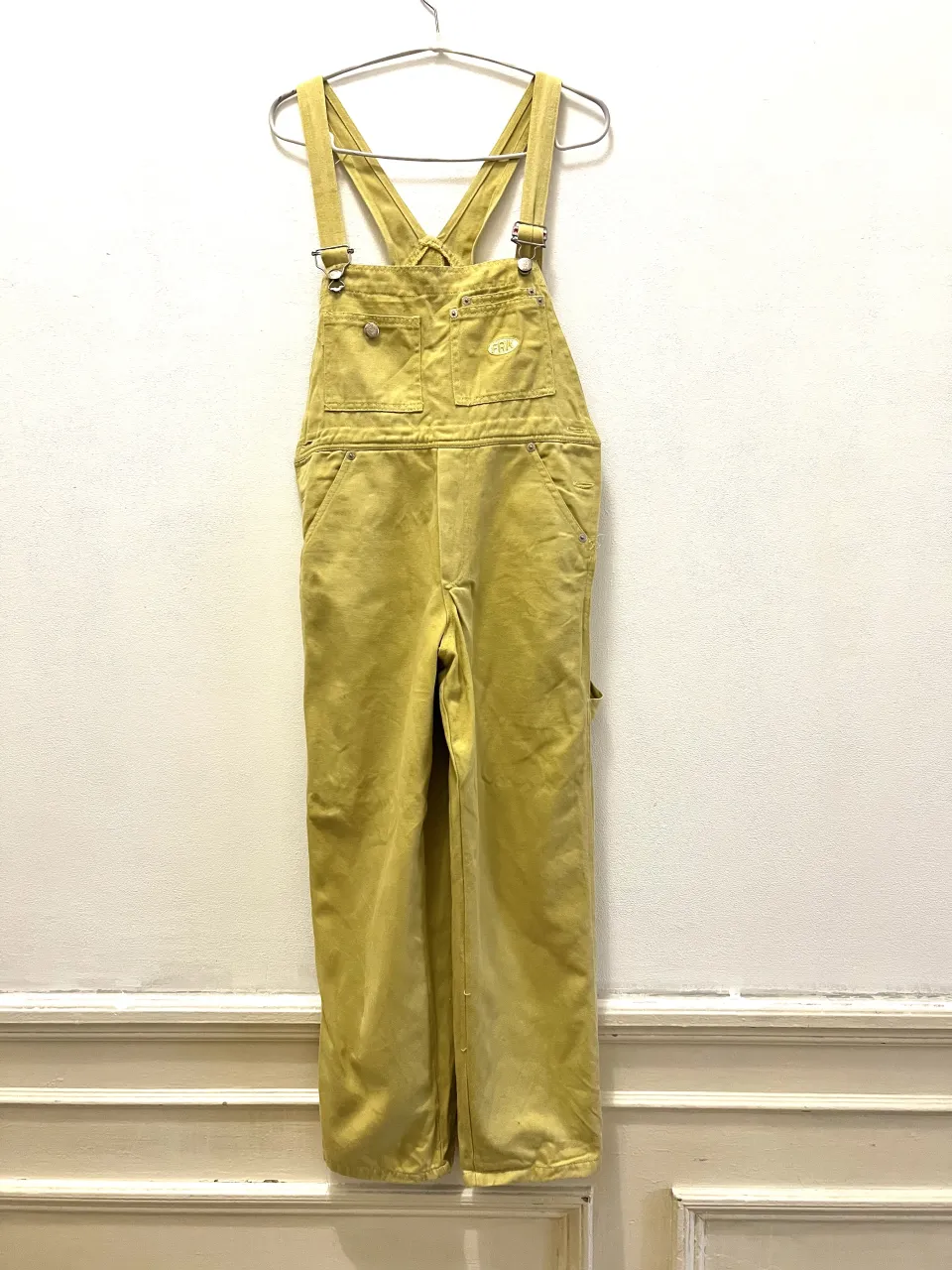 Overol de gabardina color amarillo verdoso. Posee tiradores regulables con hebillas metálicas, dos bolsillos superiores con botón y un bolsillo lateral en la pierna derecha. Cierre de botón a los costados de la cintura.