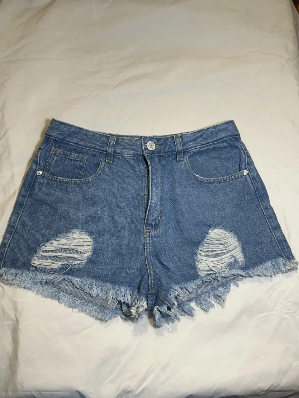 Short de jean azul (tiro medio) con roturas y flecos en el borde. Ideal para un look casual y moderno.