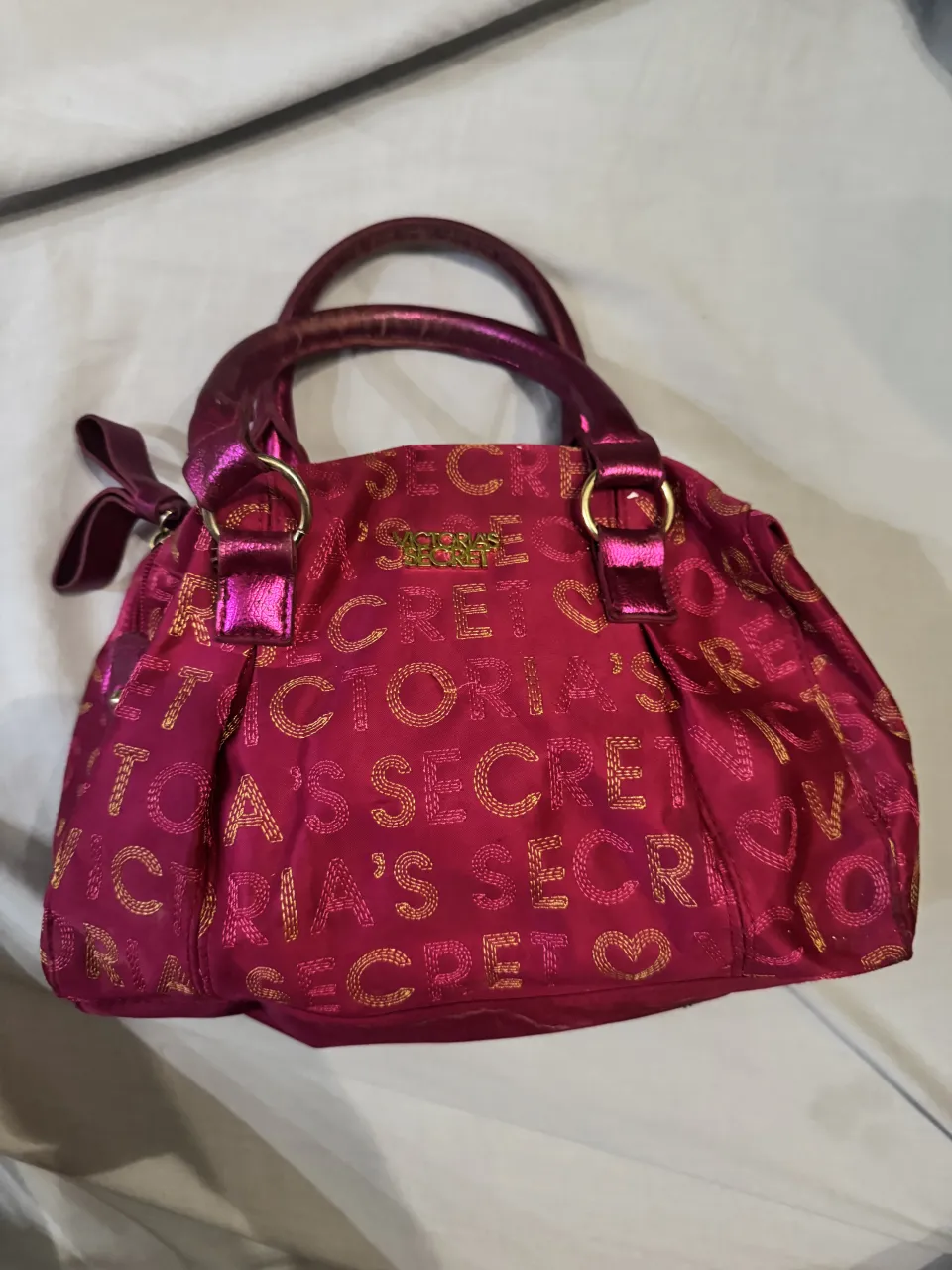 Bolso de mano color fucsia con estampa integral de letras "Victoria's Secret" en dorado. Posee dos asas cortas y un detalle de moño en uno de los laterales.