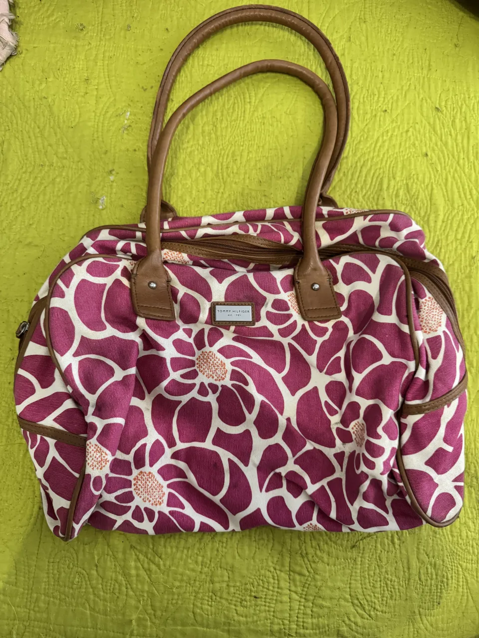 Bolso de mano con estampa floral en tonos fucsia y blanco. Posee manijas de cuero marrón y detalles de cuero en los cierres y bordes. El interior es de tela azul.