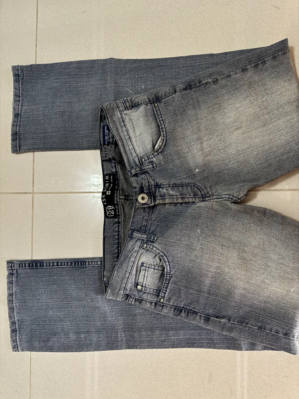 Jeans de corte slim fit, color gris desgastado con detalles de pintura blanca salpicada. Cierre de botón y cierre.