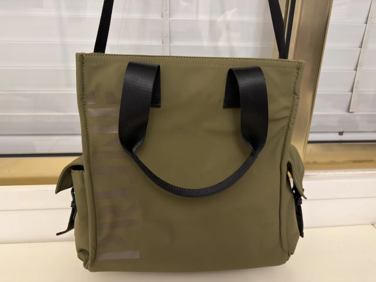 Bolso tote Prune Flight color verde musgo con dos manijas cortas negras y una correa larga. Posee bolsillos laterales con solapa y cierre superior. El interior tiene un bolsillo con cierre y dos bolsillos abiertos. Ideal para la facultad. 