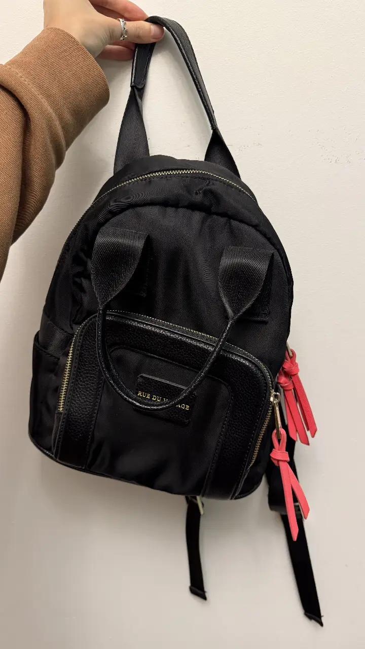 Mochila negra de tela con detalles en cuero y cierre dorado. Tiene un bolsillo frontal y detalles en rosa. Ideal para uso diario.