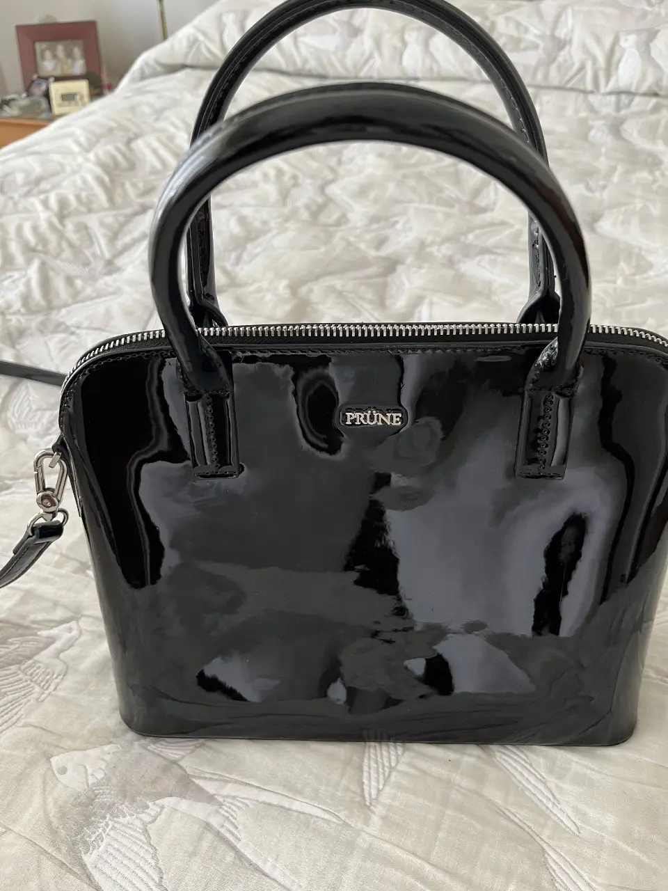 Cartera Prune negra de charol, con manijas cortas y correa larga desmontable. Ideal para llevar tus esenciales con estilo y elegancia.