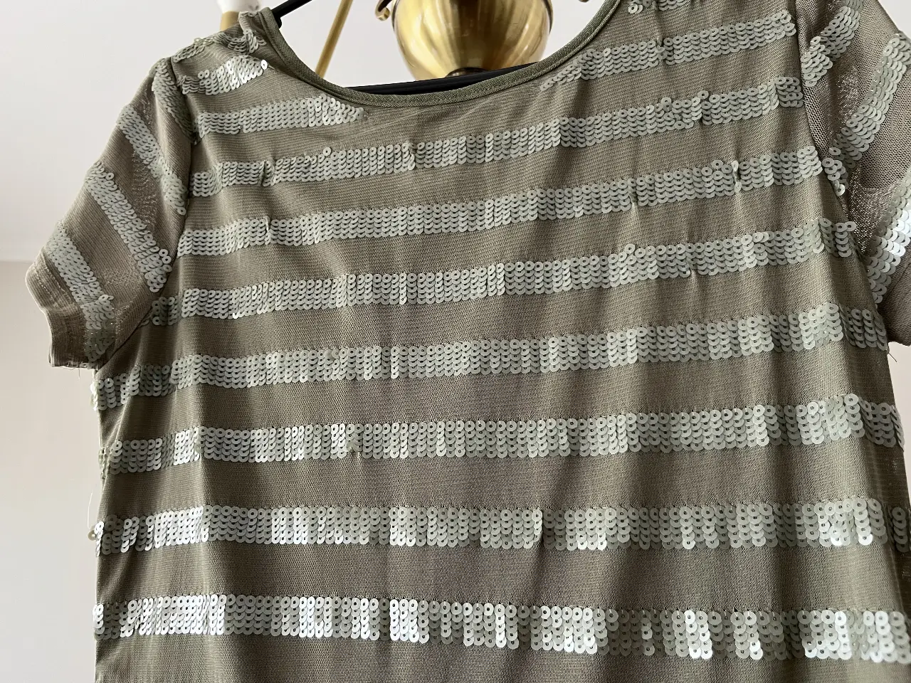 Remera de mujer con lentejuelas horizontales plateadas sobre un fondo verde oliva. Diseño elegante y moderno, ideal para un look de fiesta o salida. Marca Olga Naum.