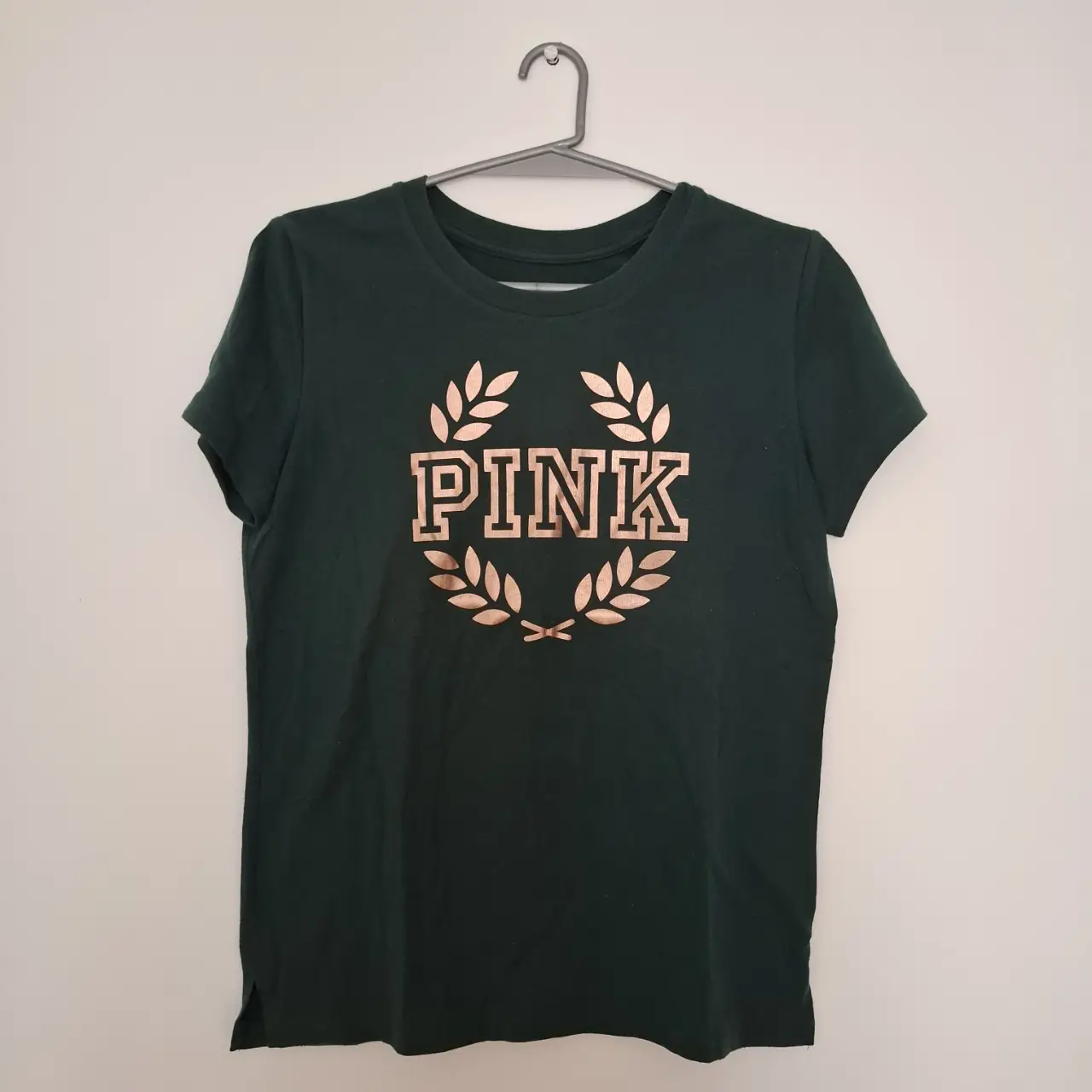 Remera Pink de Victoria's Secret, color verde oscuro con estampado "PINK" en dorado. Corte clásico, con detalle de aberturas laterales en el bajo. Ideal para un look casual y cómodo.