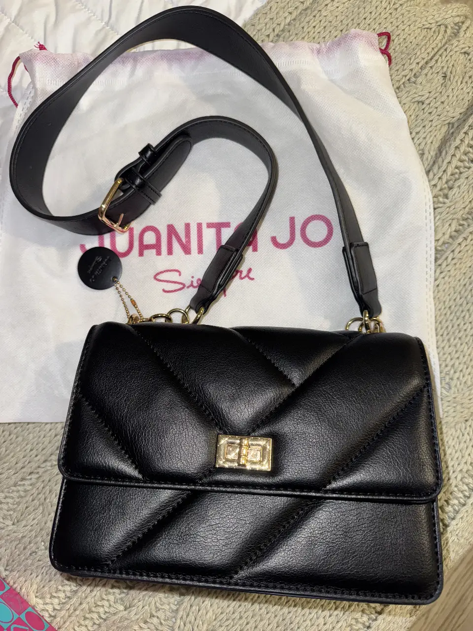 Cartera bandolera negra acolchada con cierre de solapa y detalles dorados. Incluye correa ajustable y etiqueta de la marca.