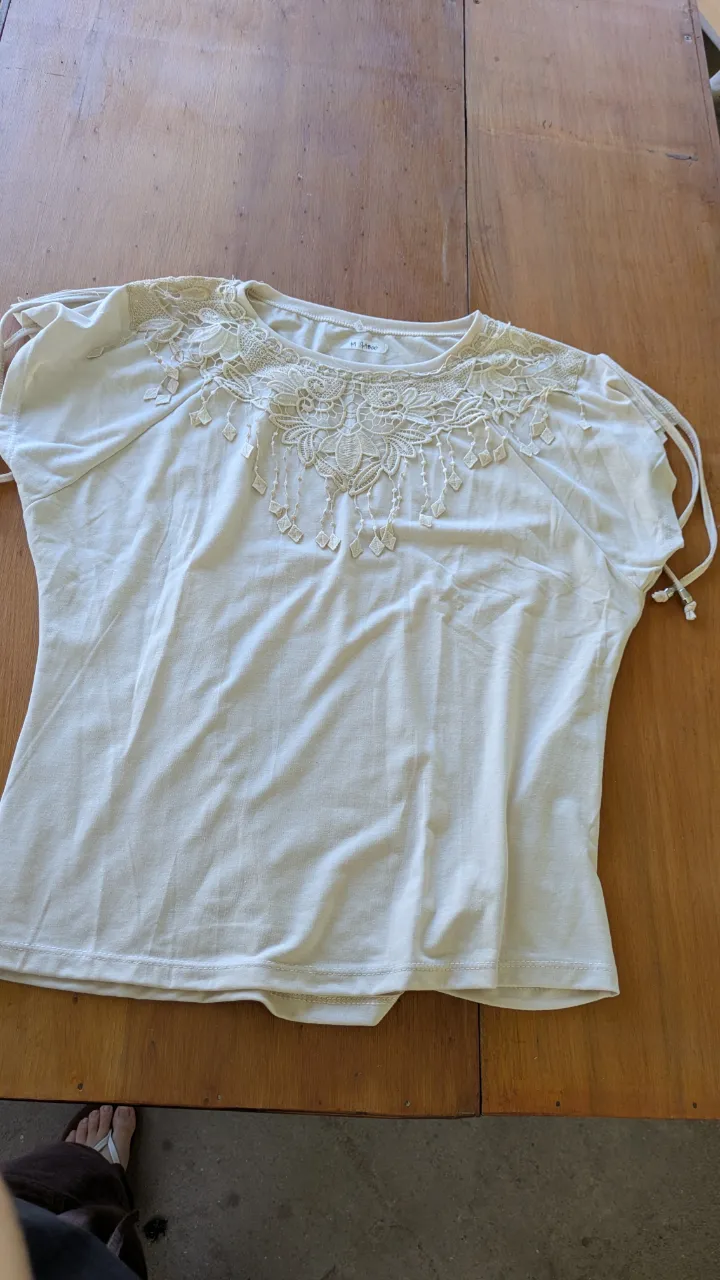 Blusa blanca con detalles de encaje y canutillos en el cuello y hombros. Mangas cortas con tiras para ajustar. Ideal para un look delicado y femenino.