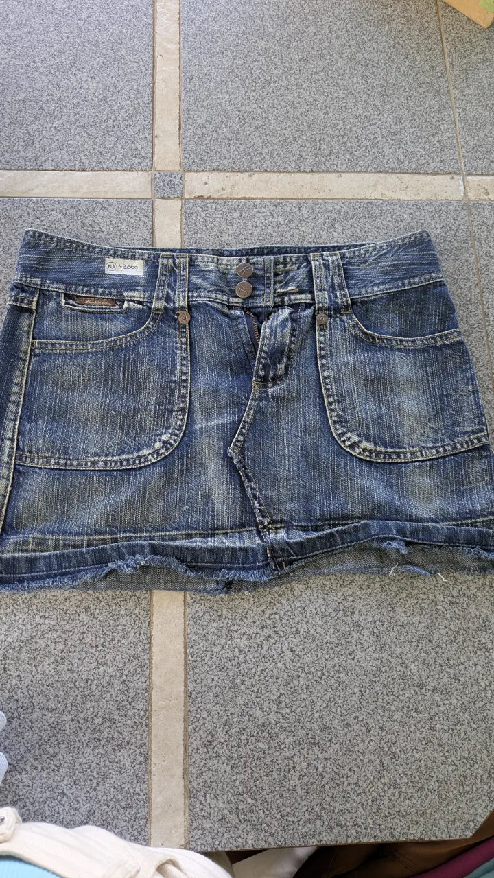 Pollera de jean azul con detalles desgastados y roturas en el borde inferior. Posee bolsillos delanteros y traseros, uno de ellos con bordado blanco.