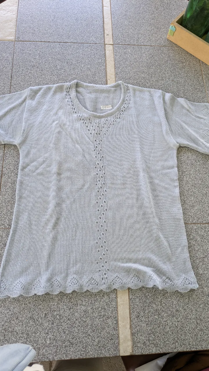 Remera de tejido fino, color gris claro, con cuello redondo y mangas cortas. Presenta un detalle de puntilla y calados en forma de V en el frente y un borde inferior ondulado.