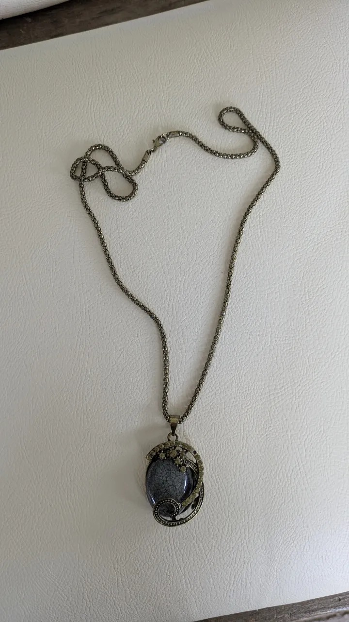Cadena con dije ovalado de piedra gris con detalles en metal con incrustaciones de strass.