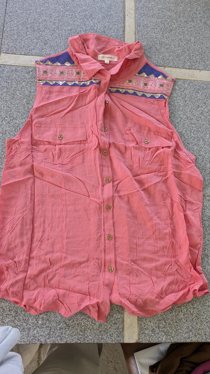 Camisa sin mangas de color rosa con detalles bordados y lentejuelas en la zona del cuello y hombros. Posee dos bolsillos en el frente y botones dorados.