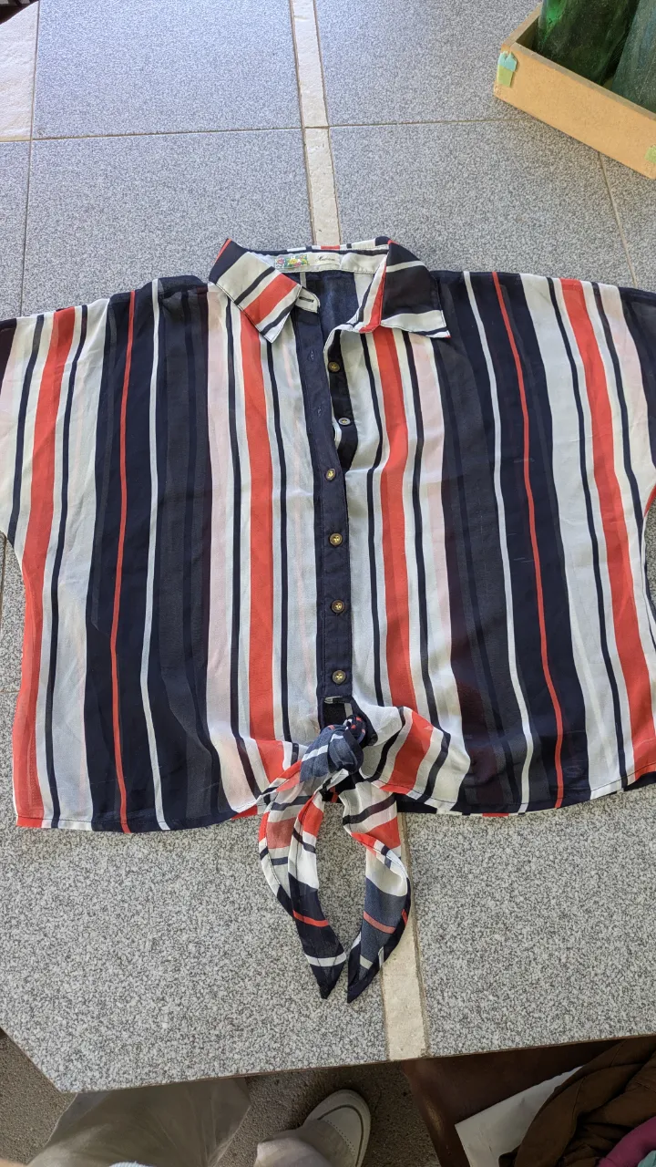 Camisa a rayas verticales en blanco, rojo y azul marino. Posee botones al frente y un nudo en la parte inferior.