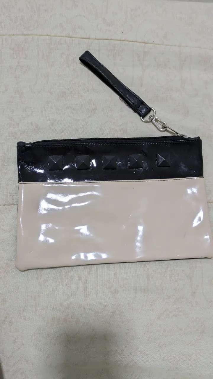 Clutch de mano para mujer, con diseño bicolor en charol. La parte superior es de color negro con detalles de tachas, y la inferior en color crudo. Incluye correa de mano desmontable.
