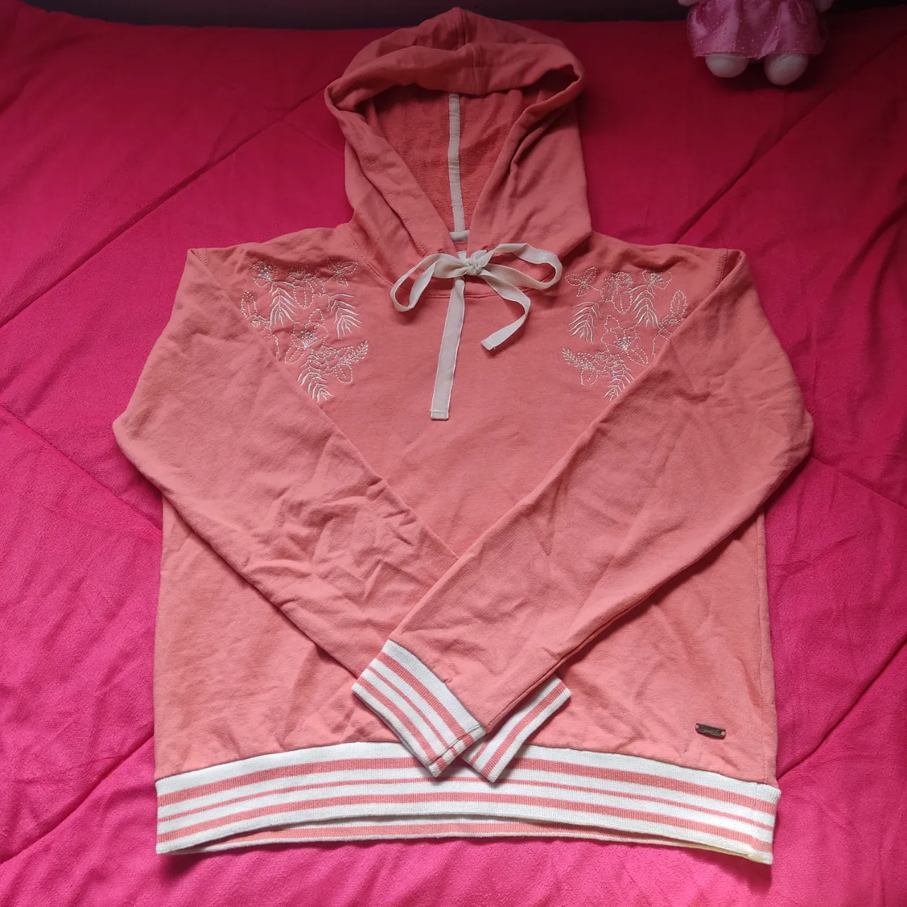 Buzo canguro de mujer con detalles bordados en blanco en la parte delantera. Tiene capucha con cinta para ajustar y puños con rayas blancas. La etiqueta indica que es de algodón y lycra. Tiene un solo uso