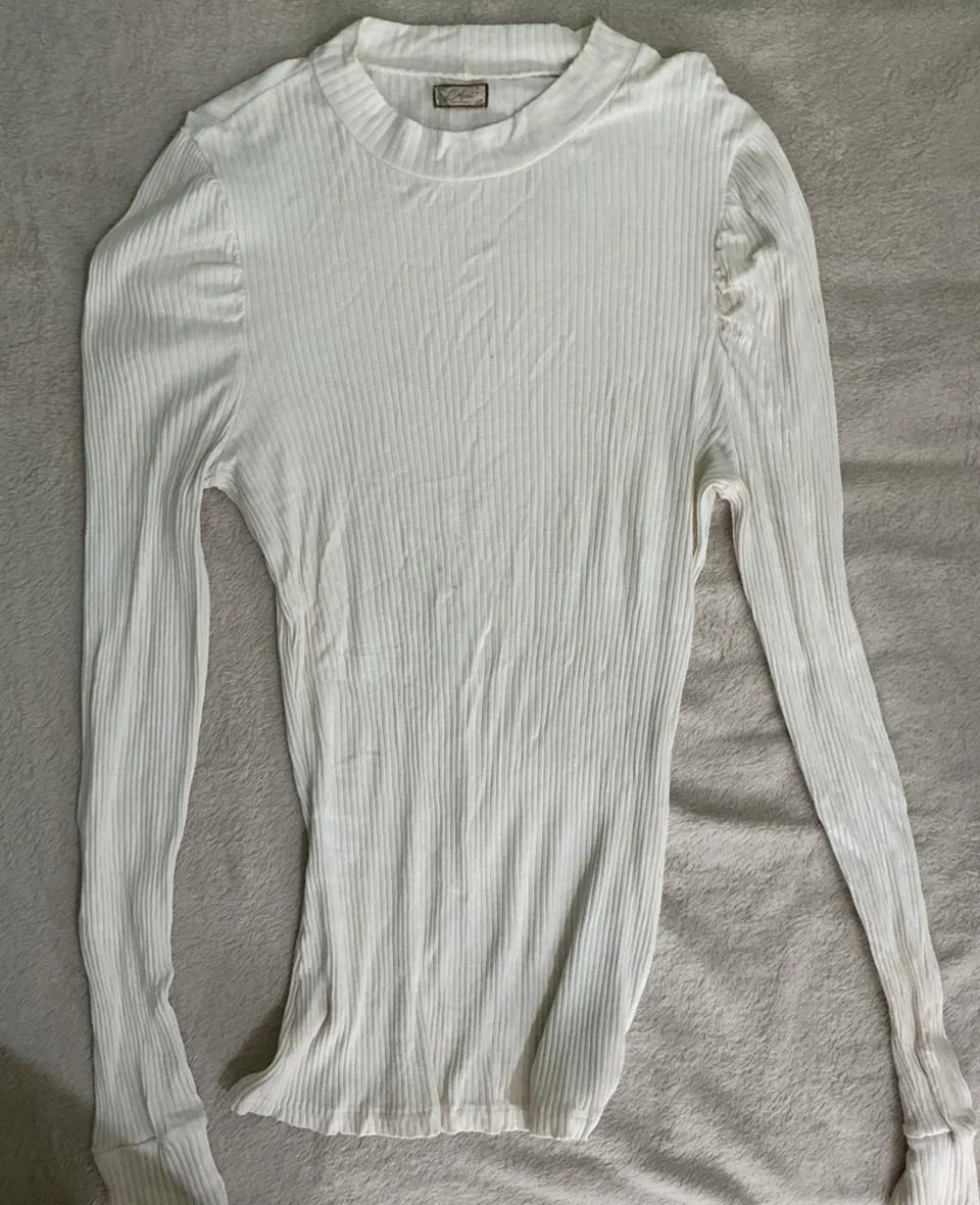 Remera blanca de manga larga, con cuello redondo y textura acanalada. Posee un detalle sutil de fruncido en los hombros que le da un toque delicado. Perfecta para un look casual y elegante.