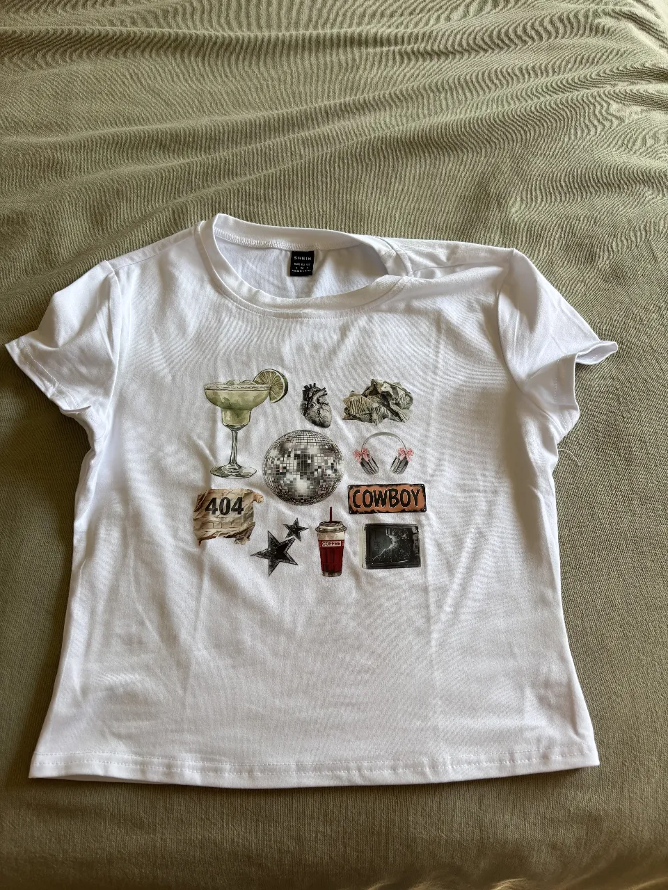Remera blanca con estampa de stickers retro y modernos. Incluye un margarita, un corazón anatómico, una bola de disco, auriculares, un cartel de 'Cowboy', un cartel de '404', una estrella negra, un vaso de bebida y una imagen de cine. Ideal para un look canchero y original.
