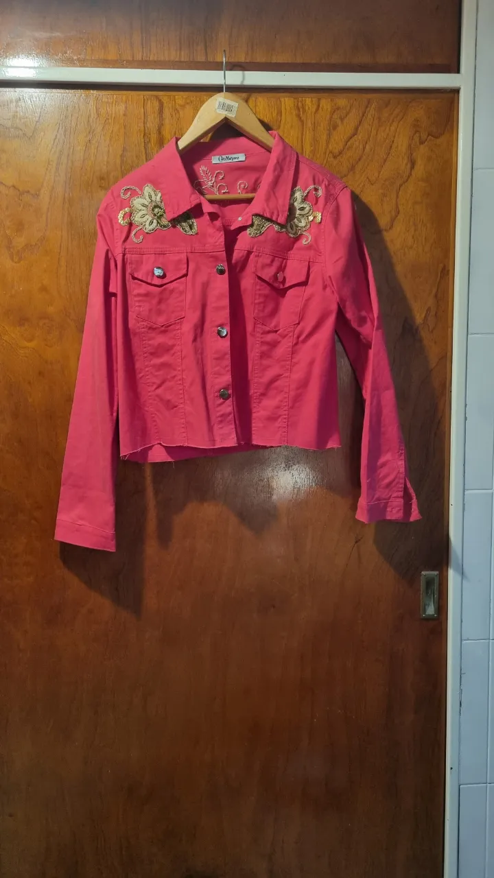 Campera corta de jean color fucsia con bordados florales y detalles de lentejuelas y mostacillas en la parte delantera y trasera. Posee cuello camisero, botones metálicos y dos bolsillos con solapa en el frente.