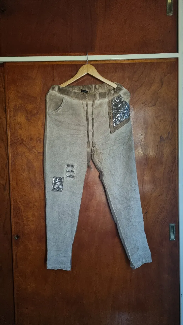Pantalón de tela color beige con detalles de lentejuelas plateadas en forma de parches y tiras. Posee cordón de ajuste en la cintura y bolsillos.