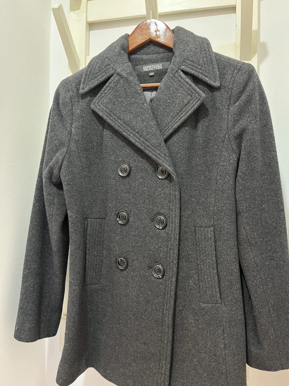 Abrigo de paño gris oscuro, doble abotonadura con cuatro bot ones visibles y dos ocultos. Cuello solapa ancho con pespuntes. Dos bolsillos frontales con tapa y pespuntes. Mangas largas.