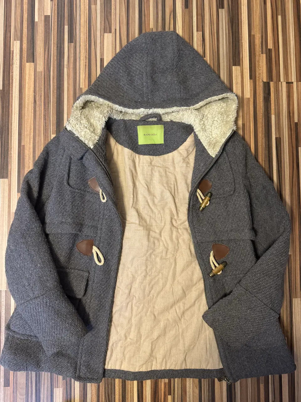 Campera de paño gris con capucha y forro de piel sintética cruda. Detalles de botones de madera y cierre frontal. Ideal para el invierno.