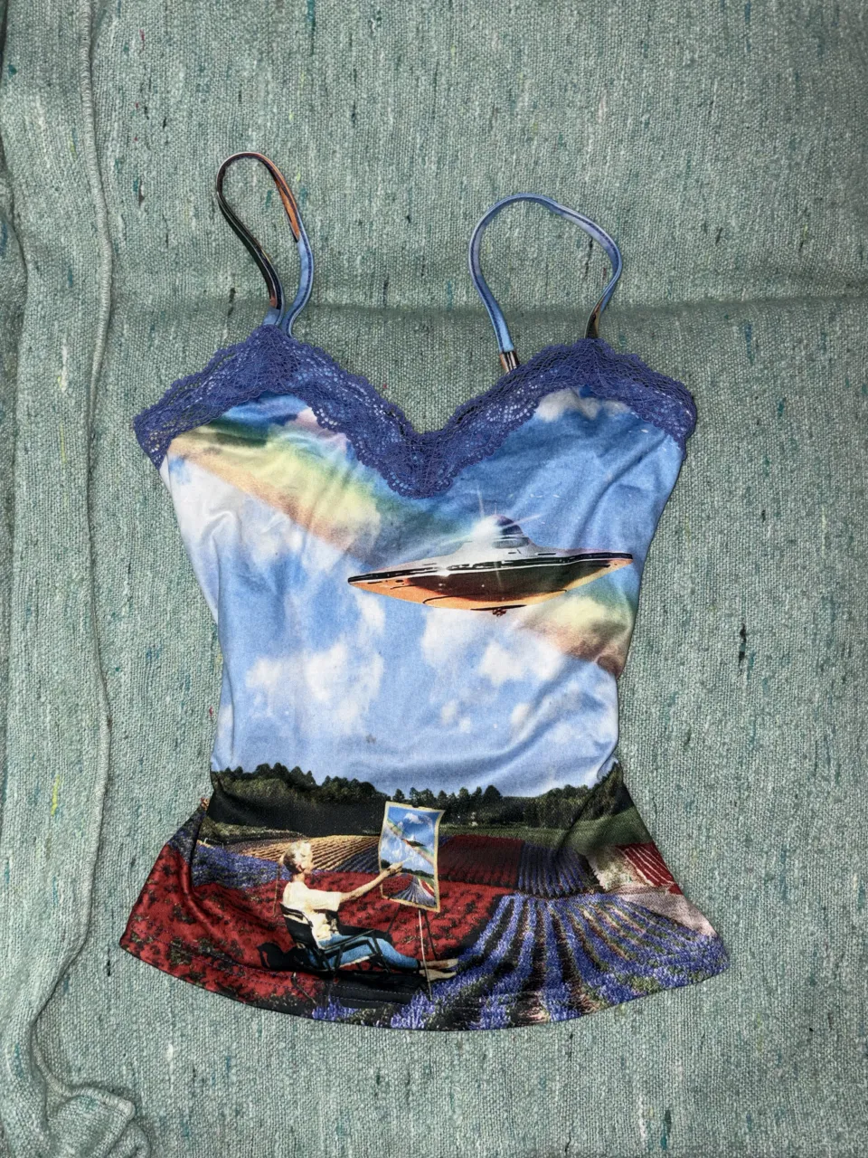 Musculosa con estampa de paisaje, cielo con ovni y figura humana pintando en un campo de flores. Posee detalles de encaje en el escote.