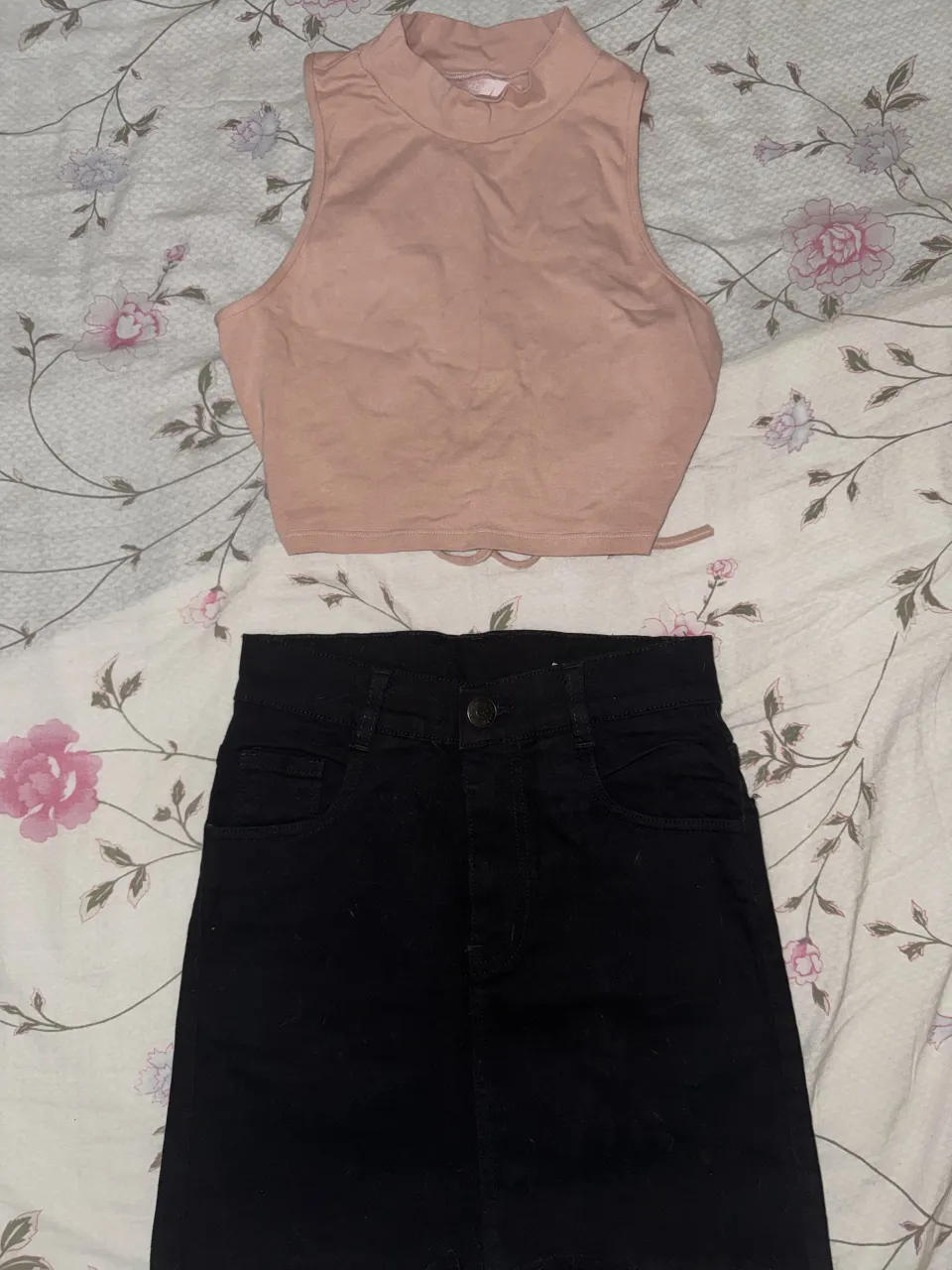 Conjunto de top y pollera tiro alto. El top es color rosa pálido, sin mangas, con cuello alto y detalle de amarre en la cintura. La pollera es negra, tiro alto, con bolsillos y cierre.