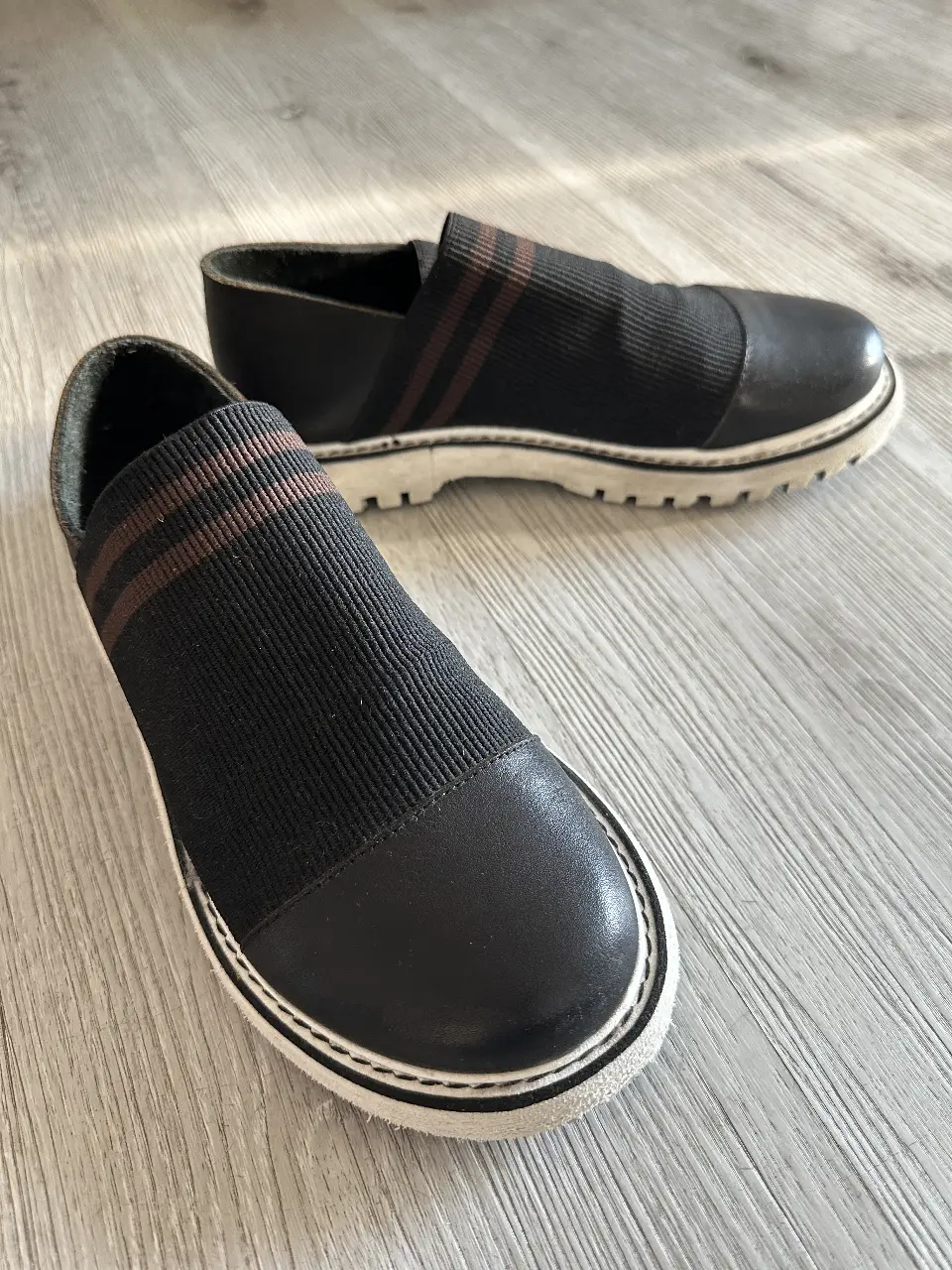 Zapatillas slip-on negras con detalles en marrón y suela blanca. Diseño moderno y cómodo, ideales para un look casual.