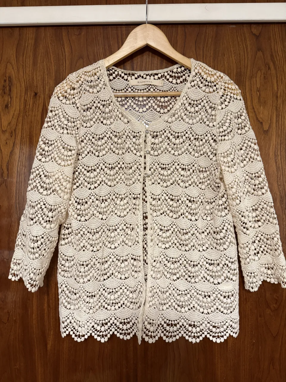 Cardigan de encaje color crudo con botones al frente y mangas 3/4. Posee un delicado diseño de ondas y pompones en el borde inferior.