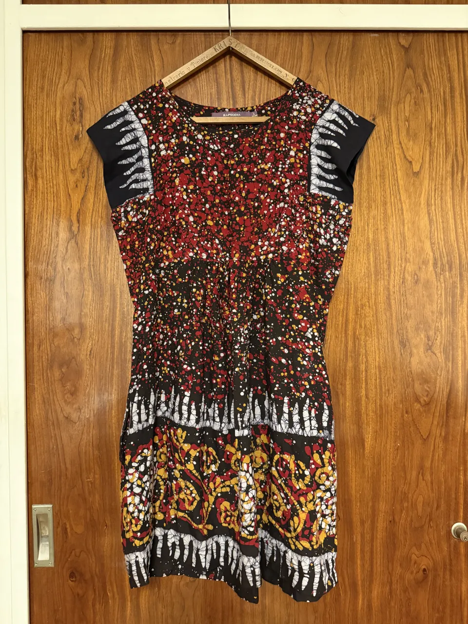 Vestido corto estampado con mangas cortas y bolsillos laterales. Presenta un diseño vibrante con salpicaduras de colores rojo, amarillo y blanco sobre fondo negro, y detalles geométricos en la parte inferior. Posee bolsillos a ambos lados