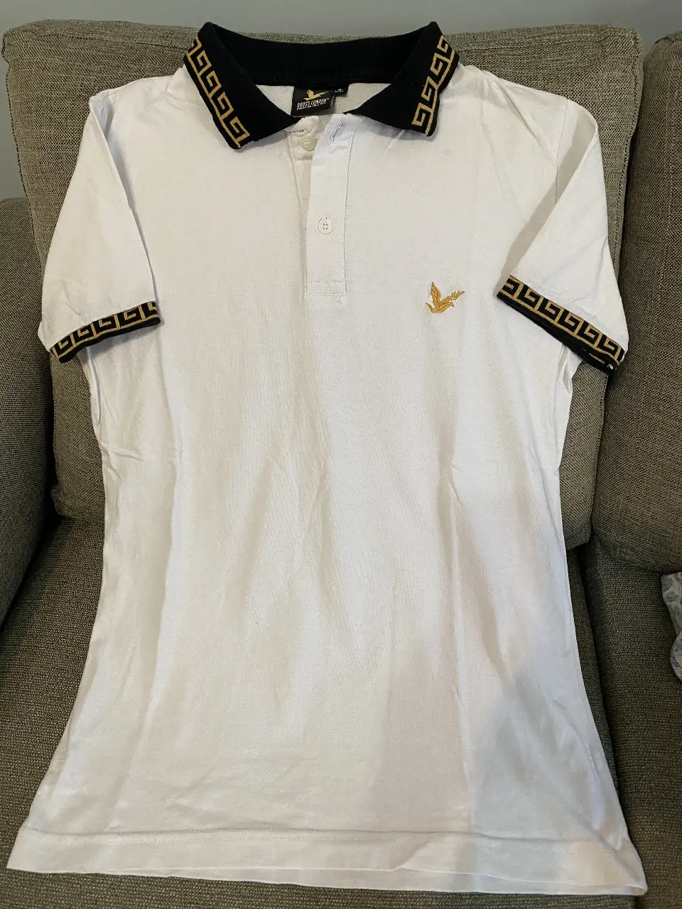 Remera polo blanca con detalles dorados y negros en cuello y mangas. Logo de ave dorado en el pecho. Ideal para un look casual y elegante.