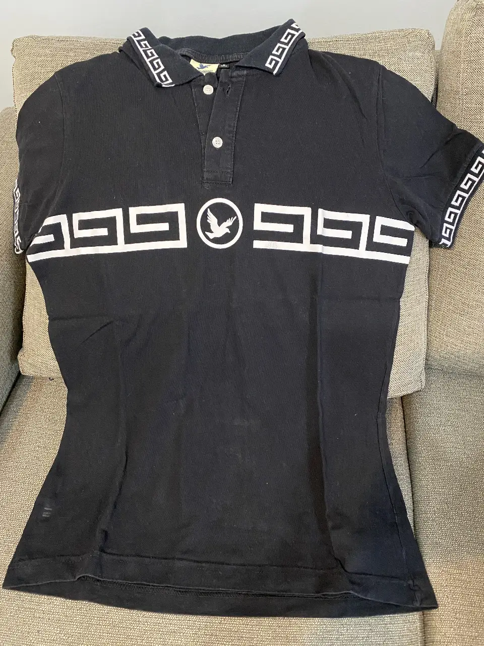 Remera polo negra con detalles geométricos blancos en cuello, mangas y pecho. Diseño con logo de ave en el centro. Ideal para un look casual y moderno.