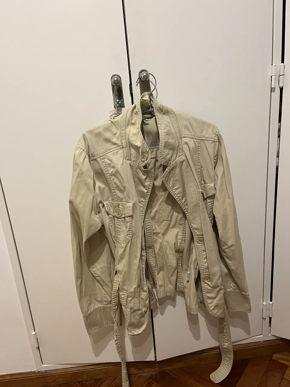 Chaqueta color crudo con cinturón de tela. Posee bolsillos con tapa y botones en el frente y puños. Cierre central.