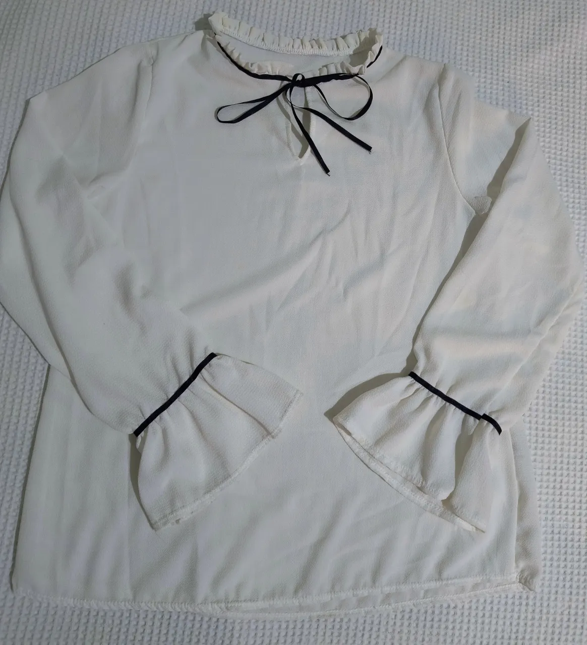 Blusa blanca de manga larga con detalles en negro. Posee cuello mao con lazo negro y mangas acampanadas con puño y detalle de volado. Ideal para un look elegante y femenino.