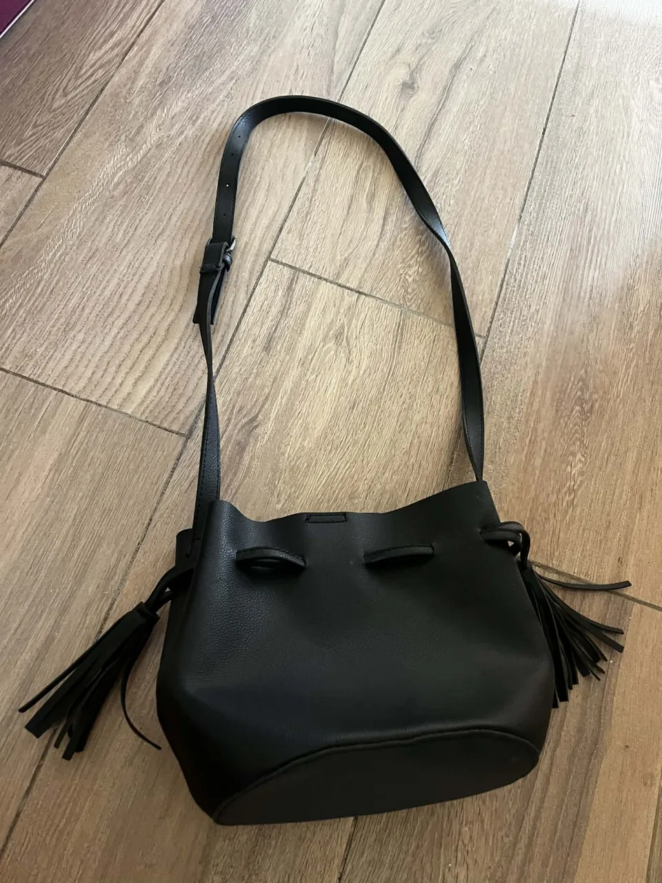 Bolso negro tipo "bucket bag" con tiras y flecos decorativos. Cierre con iman