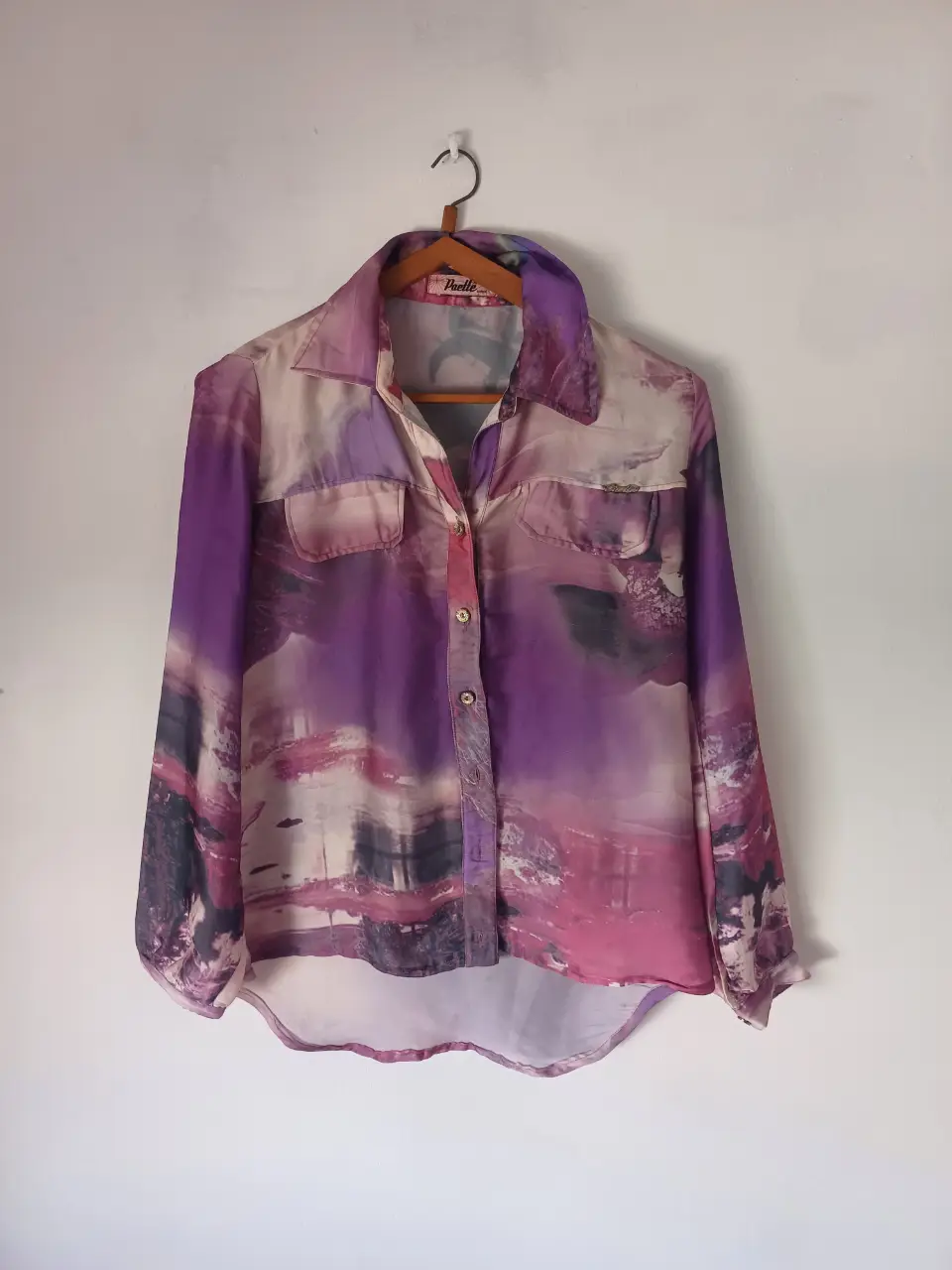 Blusa de mujer con estampado abstracto en tonos violetas y rosas. Diseño con cuello camisero, dos bolsillos delanteros y mangas largas.
