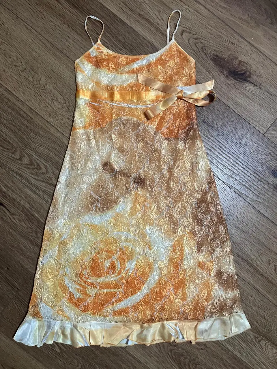 Vestido mini de fiesta con tirantes finos y detalle de moño en la cintura. Diseño con estampado floral en tonos dorados y naranjas, ideal para eventos especiales.