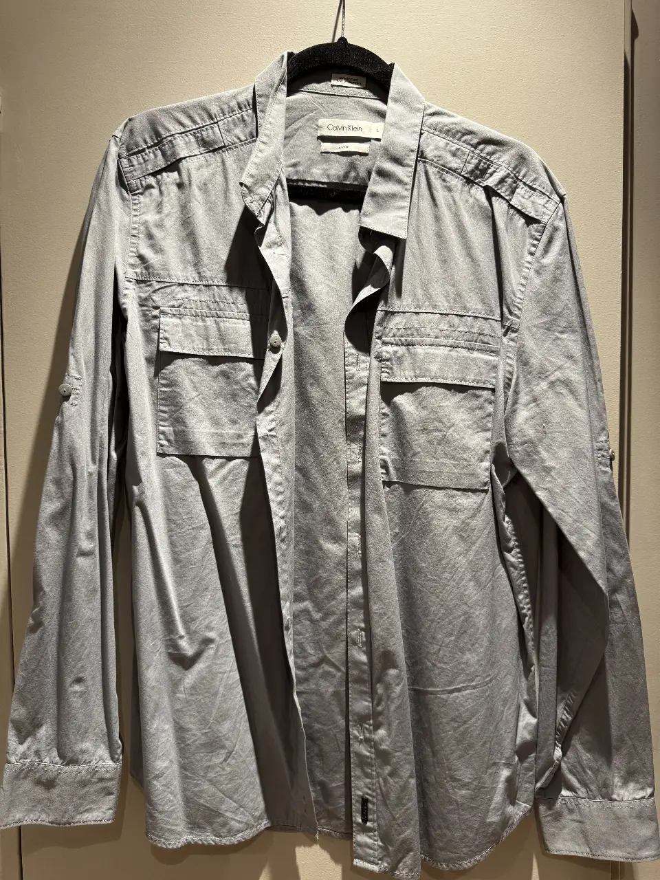 Camisa de hombre Calvin Klein, manga larga, color gris, con dos bolsillos frontales y detalles en los hombros. Talle L, corte slim fit. Ideal para un look casual y moderno.