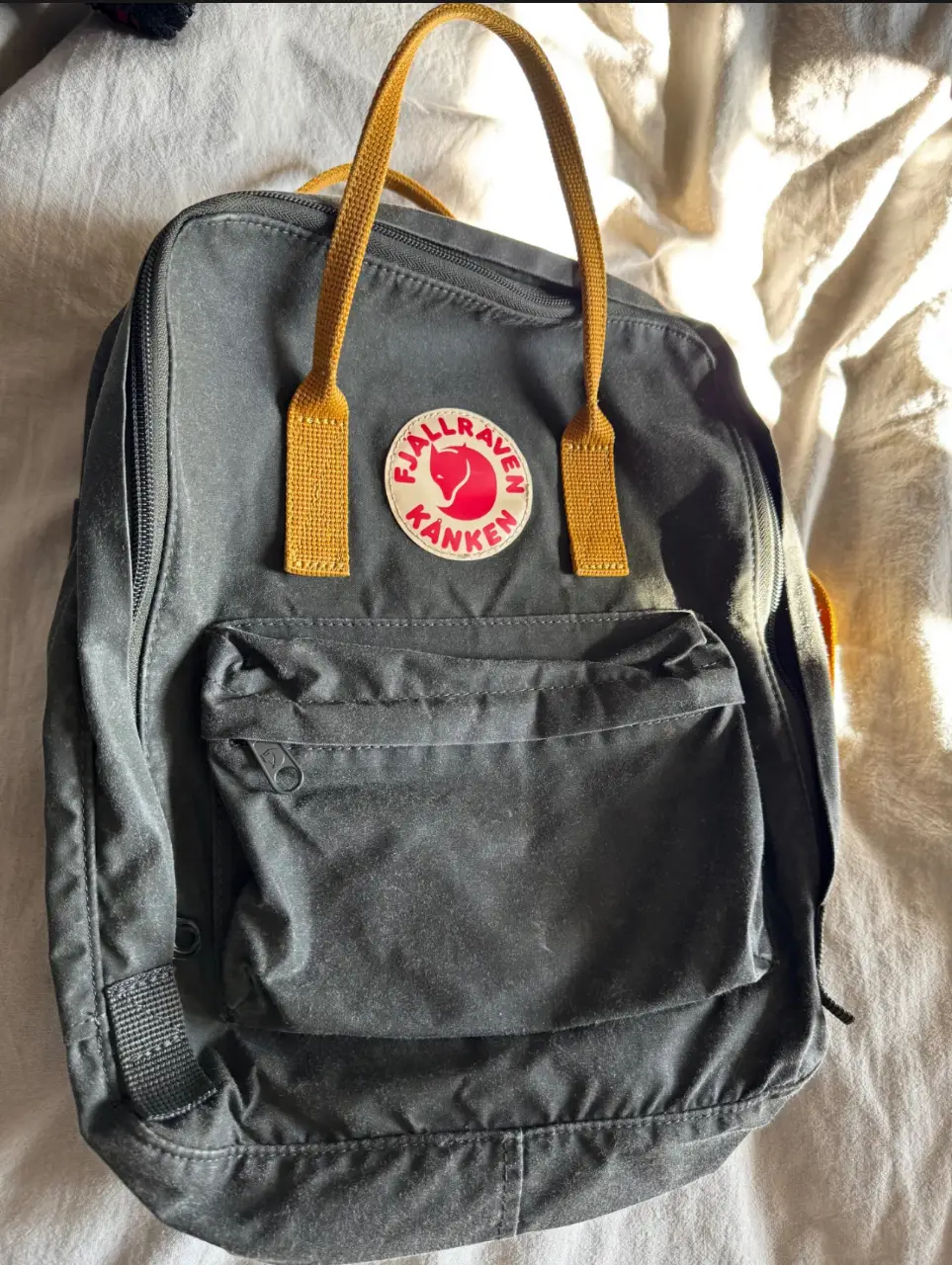 Mochila Fjallraven Kanken en color gris con detalles en amarillo. Ideal para uso diario, con un diseño clásico y resistente. Cuenta con un bolsillo frontal con cierre y correas ajustables.