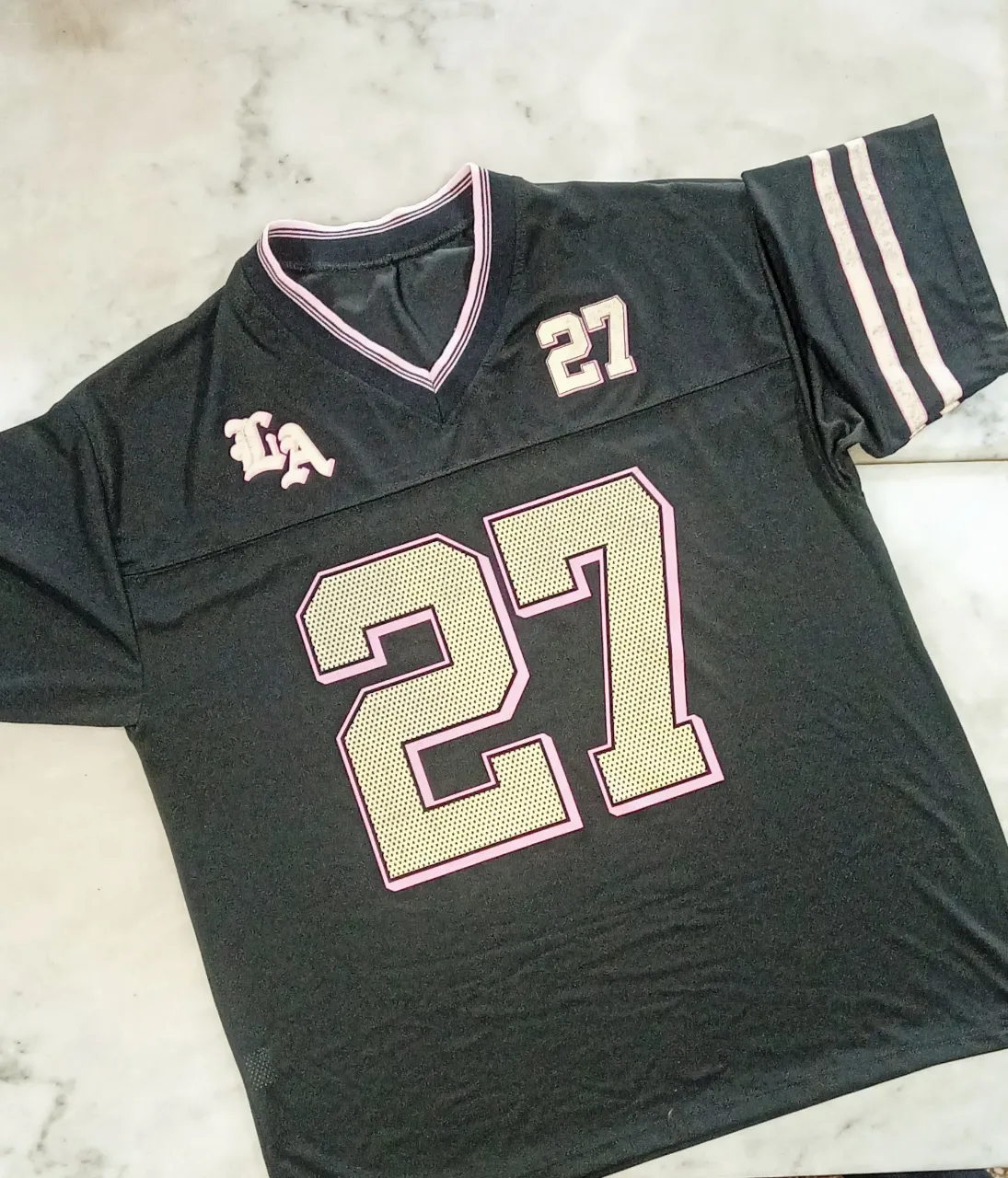 Remera deportiva negra con número 27 en dorado y rosa. Diseño inspirado en uniformes de fútbol americano, ideal para un look urbano y canchero.