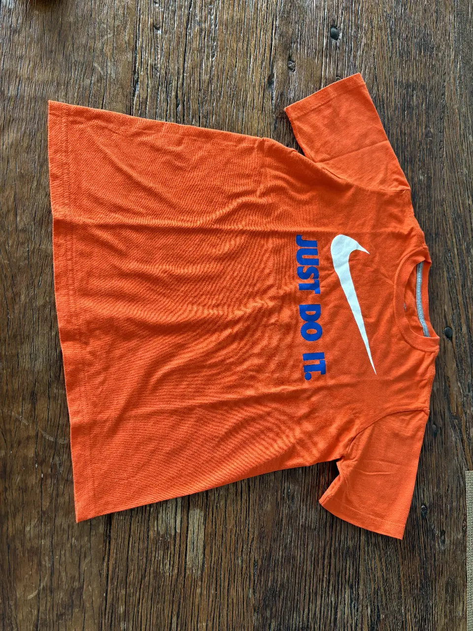 Remera deportiva naranja de la marca Nike, con el logo y la frase "JUST DO IT" en color blanco y azul. Ideal para actividades físicas.