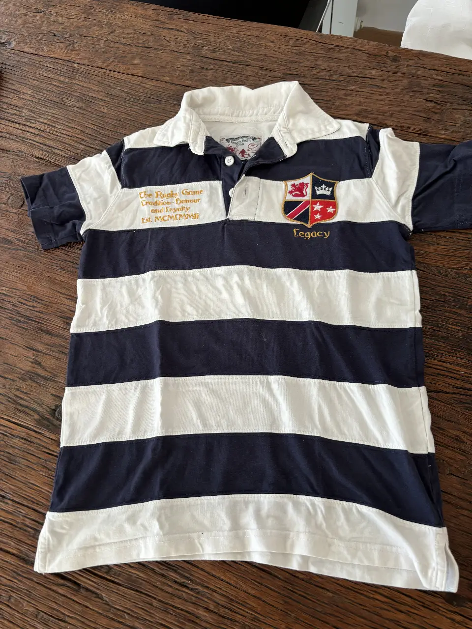 Remera de rugby a rayas azul y blanca con detalles dorados y escudo. Ideal para un look casual y deportivo.