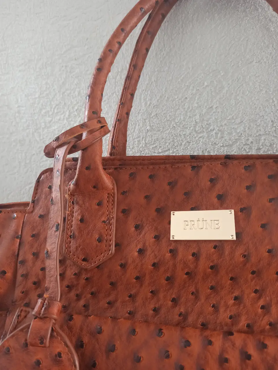 Cartera PRUNE de cuero con textura de avestruz, con manijas y detalles en cuero. Posee un aplique metálico con el nombre de la marca. Interior espacioso con bolsillo interno con cierre.
