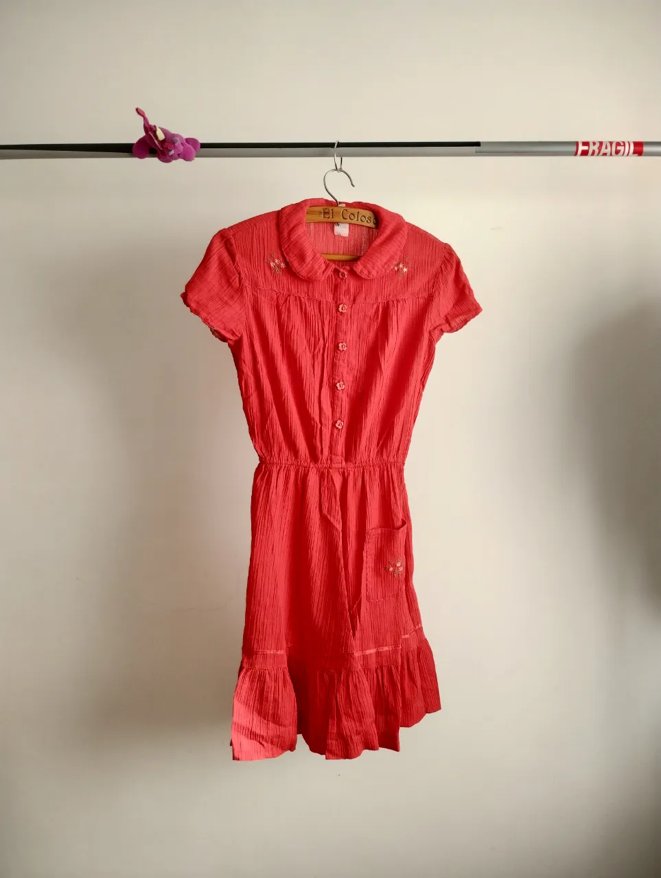 Vestido vintage rojo con detalles bordados florales en el cuello y un bolsillo. Posee cuello camisero, botones al frente y cintura elastizada. La falda tiene un corte con volados y terminación fruncida, ideal para un look fresco y femenino. Perfecto para salidas casuales o eventos de día.