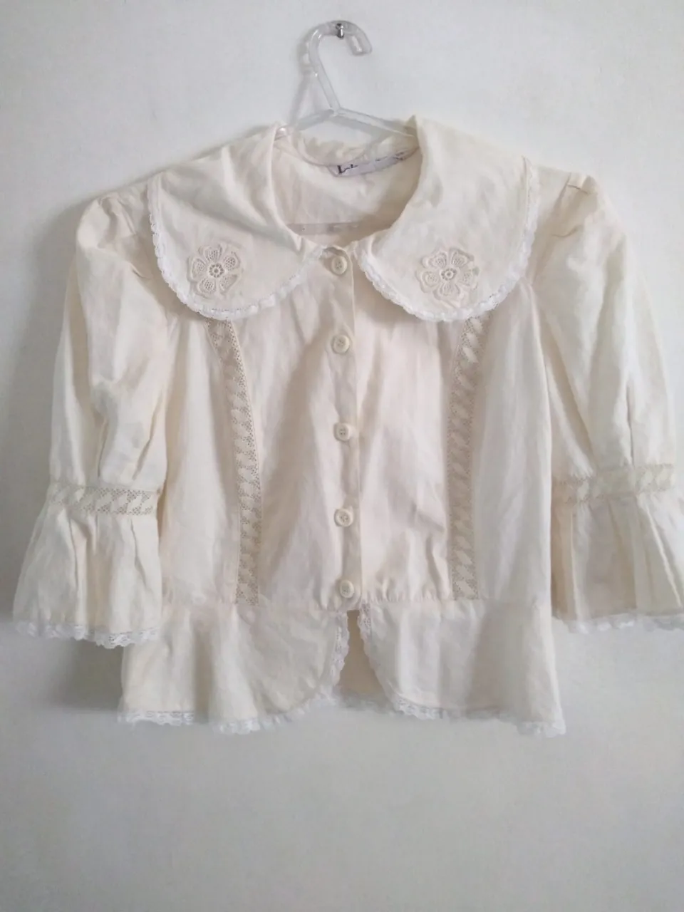Blusa marfil/crudo de mangas cortas con volados y detalles de encaje. Posee un cuello redondo con puntillas y un diseño floral bordado en la parte delantera. Los puños de las mangas también tienen volados y encaje, dándole un toque romántico y delicado. Perfecta para un look femenino y coqueto.