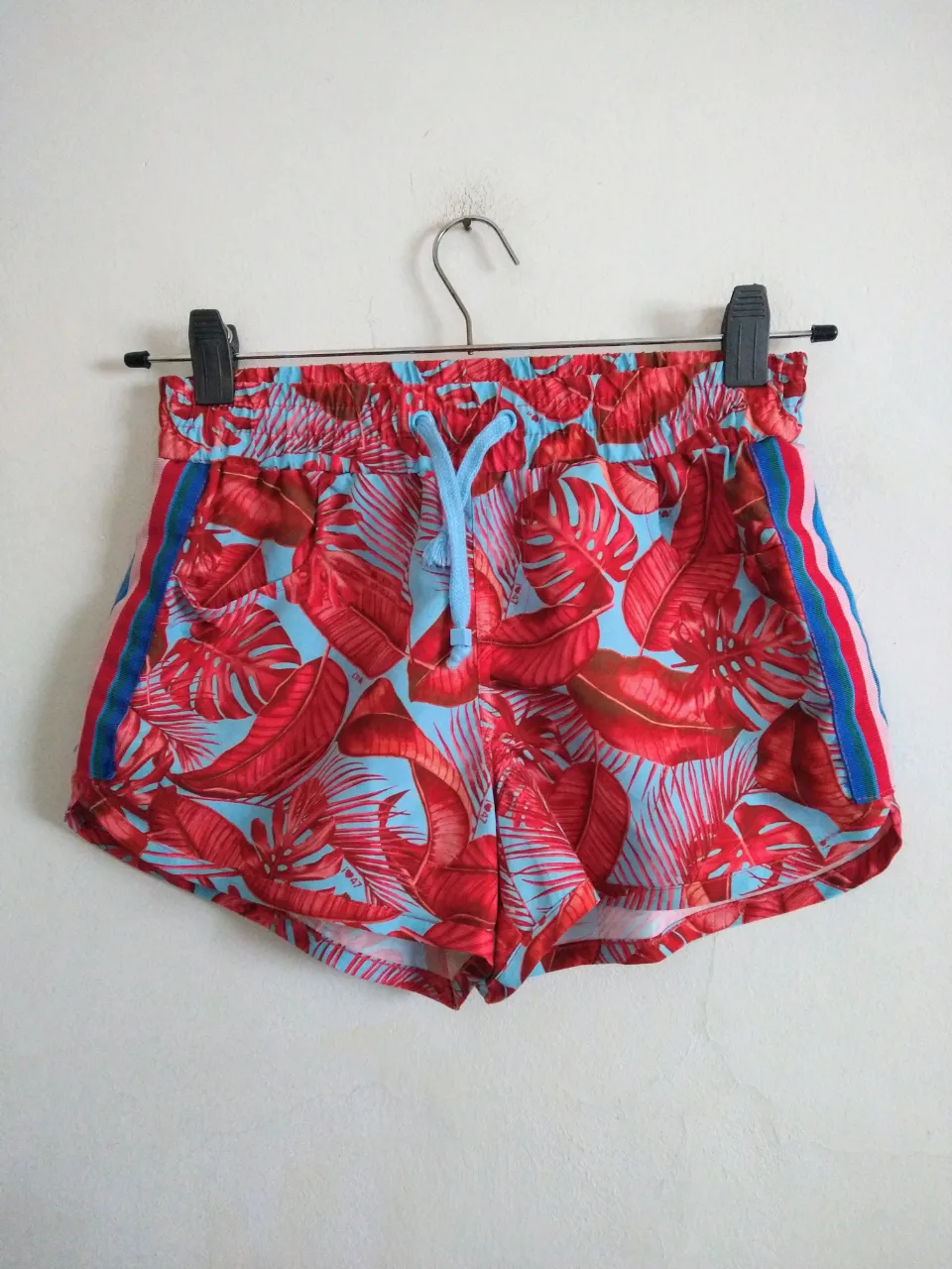 Shorts de verano con estampado tropical de hojas rojas sobre fondo celeste. Tiene bolsillos y detalles de tiras laterales en rojo, blanco y azul. Ideales para un look playero o casual.