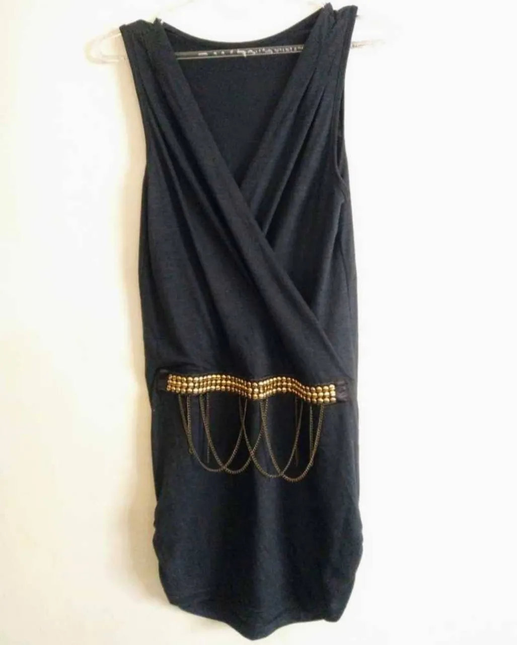 Vestido negro sin mangas, con escote cruzado y detalle de cinturón con tachas doradas y cadenas colgantes. Posee fruncido en parte de abajo que estiliza la figura. Ideal para una salida.