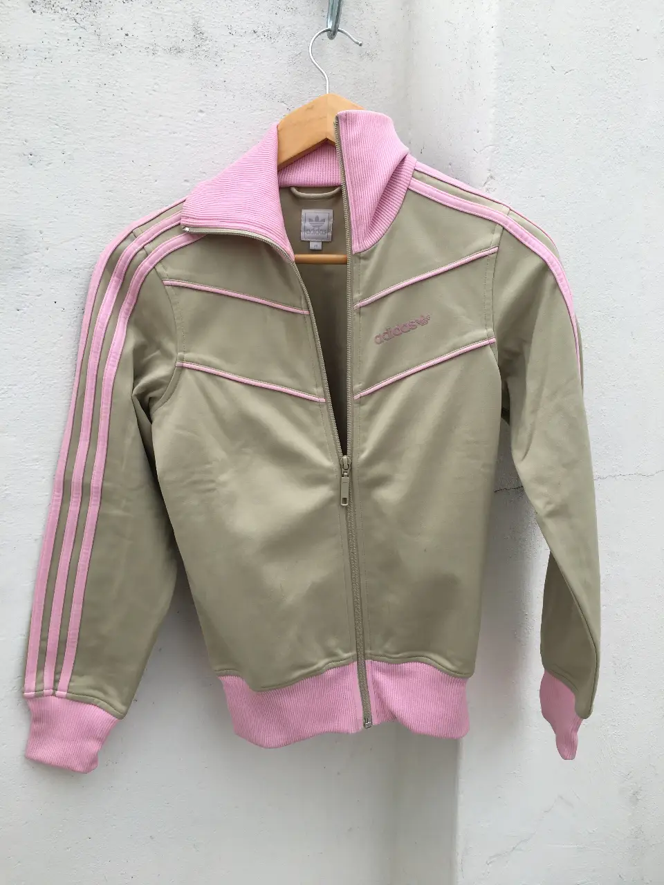 Campera deportiva Adidas de color beige con detalles en rosa. Tiene las tres tiras clásicas de la marca en las mangas y el logo bordado en el pecho. Posee cierre y puños y cuello en rosa. Ideal para un look deportivo y casual.