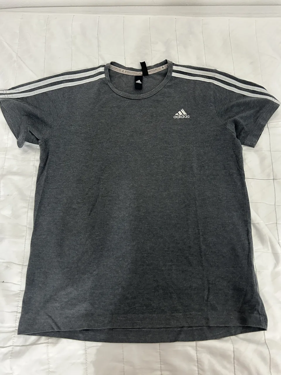 Remera gris de manga corta con las icónicas tres tiras de Adidas en los hombros. Un clásico deportivo para tu día a día.